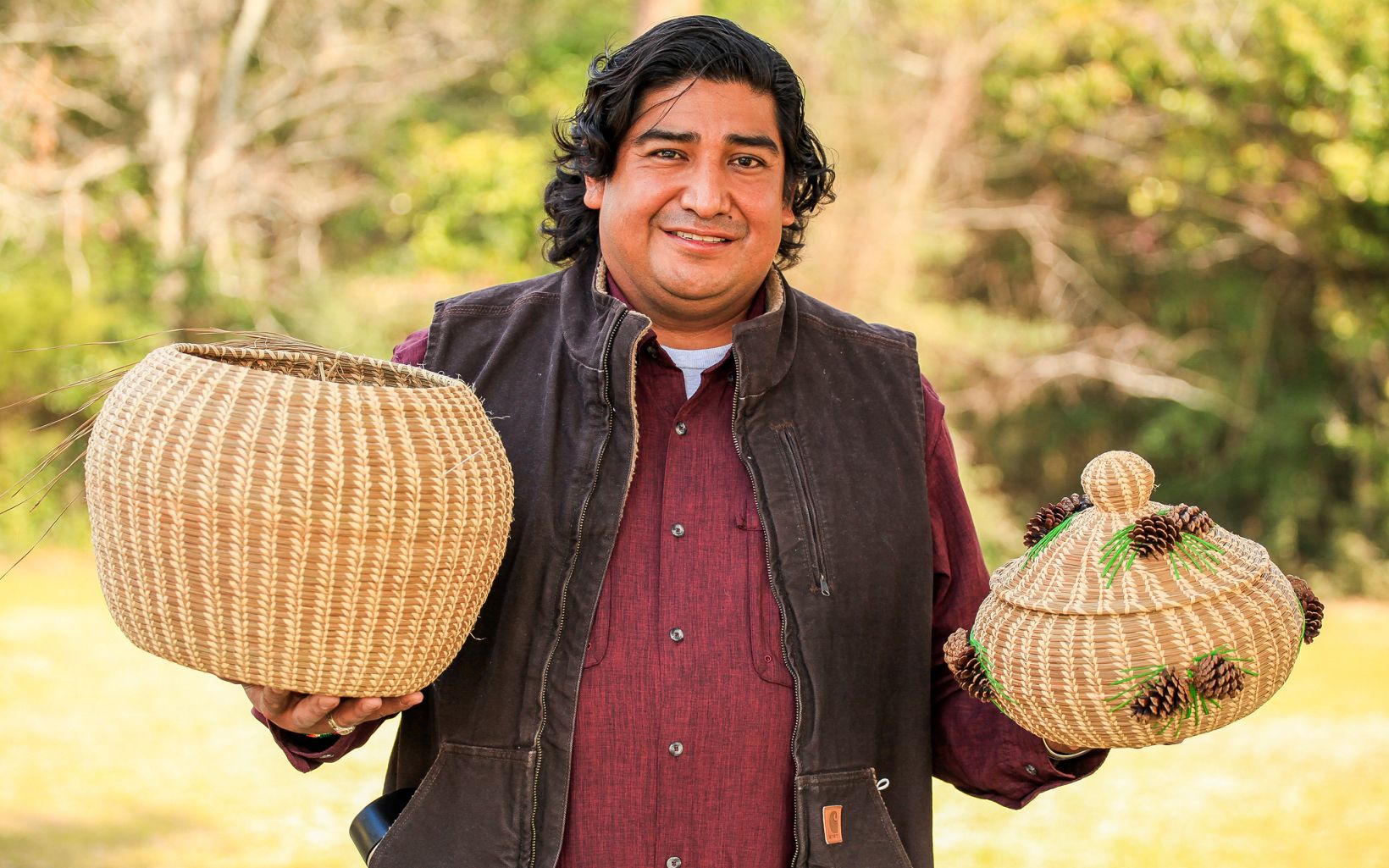 Elliott Abbey, a basket weaver of the Coushatta Tribe of Louisiana, stands outside holding brown patterned baskets, one adorned with green raffia and pinecones, in the palm of each of his hands. 