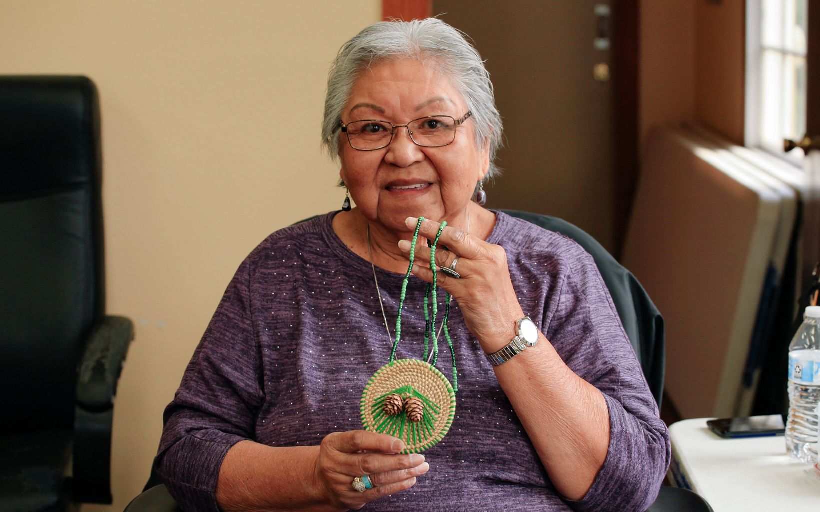 Joyce Poncho of the Alabama-Coushatta Tribe of Texas sits, holding a circular pendant necklace woven from longleaf pine needles that is decorated with green raffia, pinecones, and green beads.