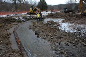 A section of stream begins to take shape in the middle of a muddy construction area. In the background a man wearing an orange hard hat stand in ankle deep water. A yellow backhoe sits on the bank.