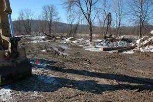 A large muddy construction area where a new stream bed will flow. A yellow earthmover is visible at the left. Logs and a large pile of rocks sit together on the right, waiting for placement.
