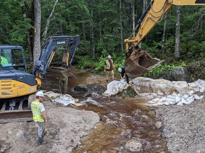 two backhoe tractors list white bags out of a stream