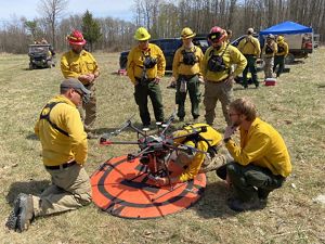Several people in yellow fire gear gather around a drone on the ground.