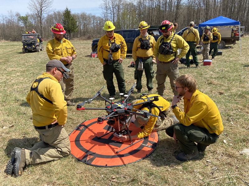 Several people in yellow fire gear gather around a drone on the ground.