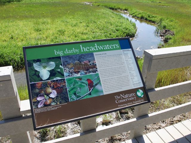 An interpretive sign attached to a wooden structure overlooks a meandering stream.