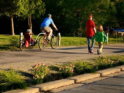 Man on a tandem bike with child in the back and woman with a second child walk along a trail bordered by planted pink flower