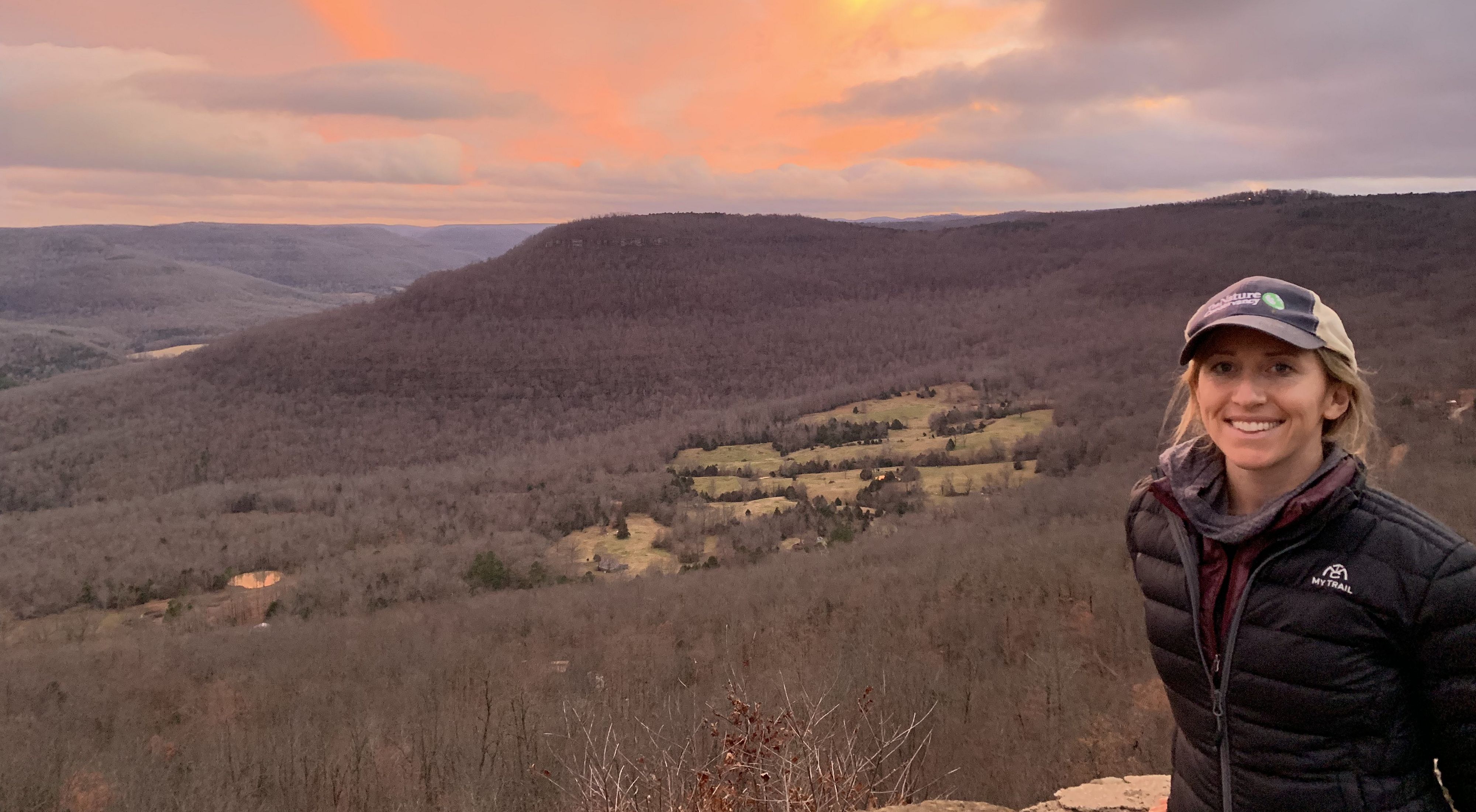 Hannah Birgé stands in front of a rock outcropping under a pinkish sunset sky.
