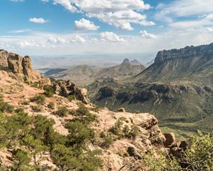 View of towering rocky mountains lined with desert shrubs and small trees.