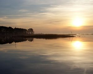 Sunset over the Blackwater National Wildlife Refuge. The sun hangs low in the sky above the horizon behind a haze of clouds. Its light is reflected in the still water of the Blackwater River.