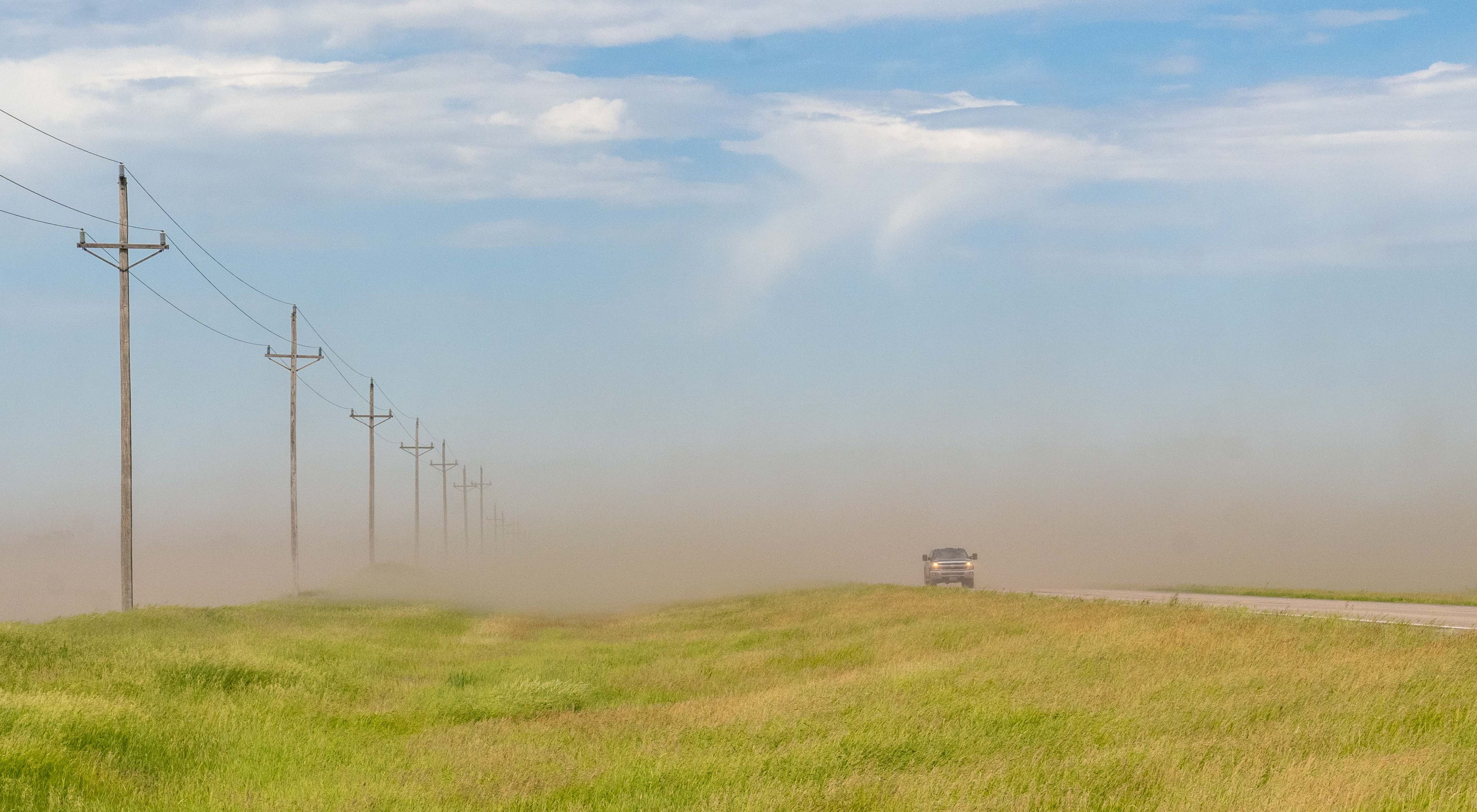 soil blowing across a field with a road and vehicle on the right.