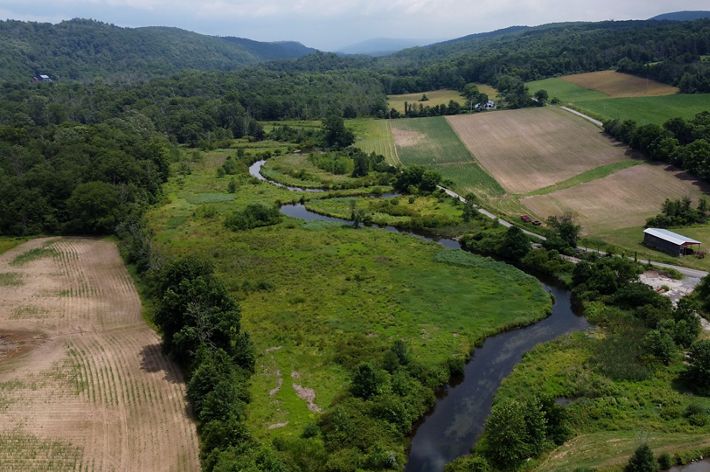An aerial view of a parcel of land containing large fields, grass and lush green trees. A winding stream flows through the center of the fields.