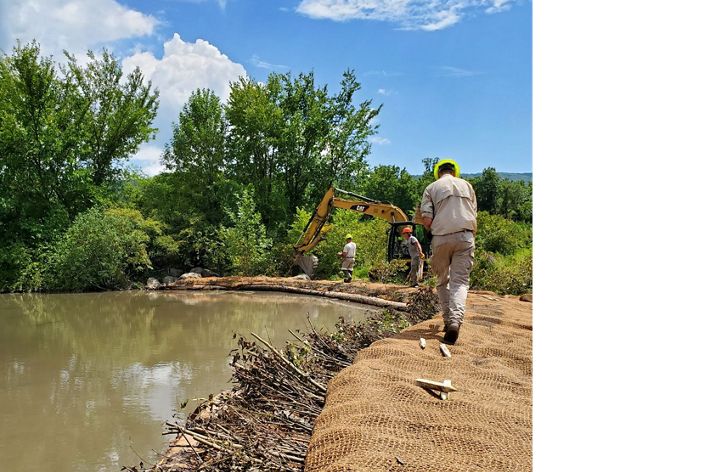 A person in construction gear walks to right side of a body of water where the surrounding land is being actively worked on by heavy machinery and construction personnel.