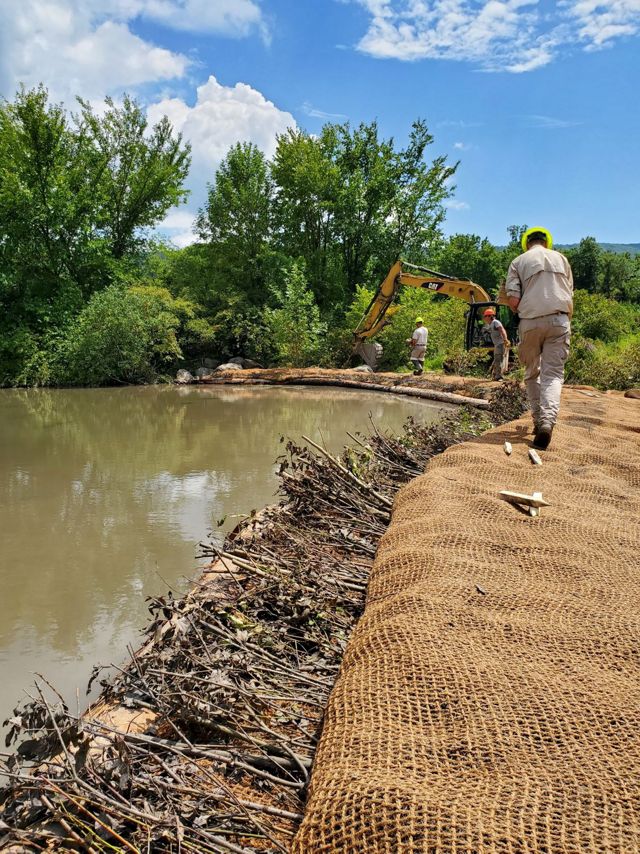 A person in construction gear walks to right side of a body of water where the surrounding land is being actively worked on by heavy machinery and construction personnel. 