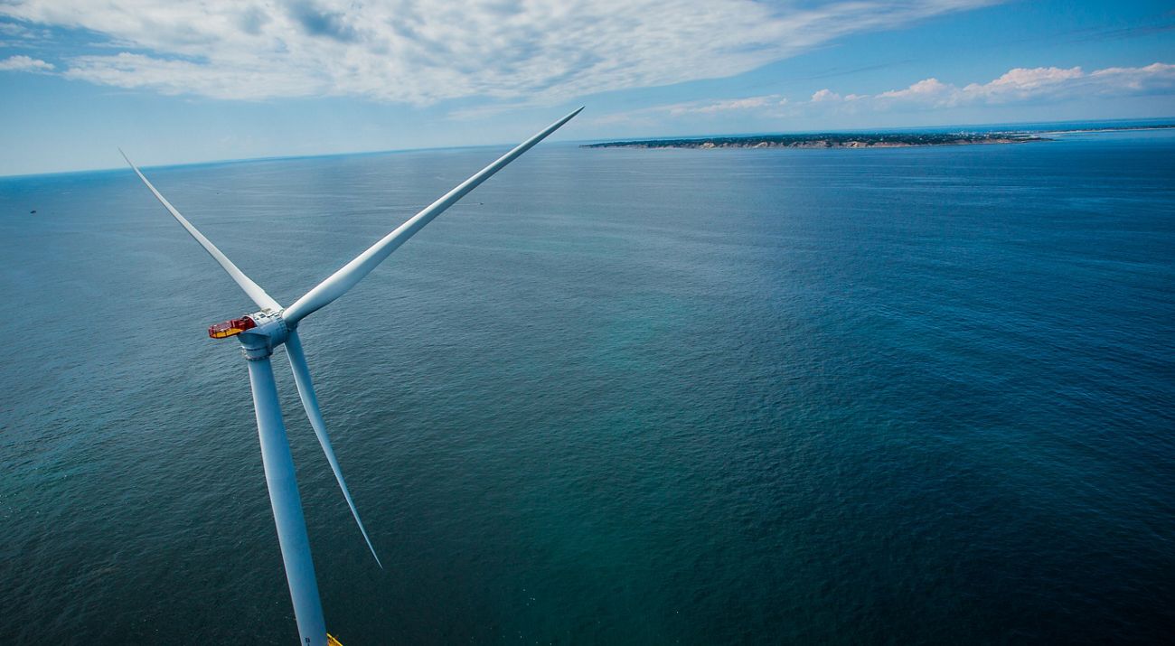 A la izquierda se muestra una turbina eólica situada en la cima de un océano abierto, con una pequeña porción de tierra con pasto verde a la vista. Se ven nubes en un cielo azul.