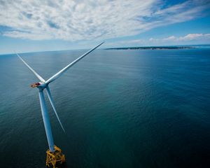 A wind turbine is pictured at left and sits on top of a wide open ocean, with a small patch of land with green grass in view. Clouds are seen on a blue sky.