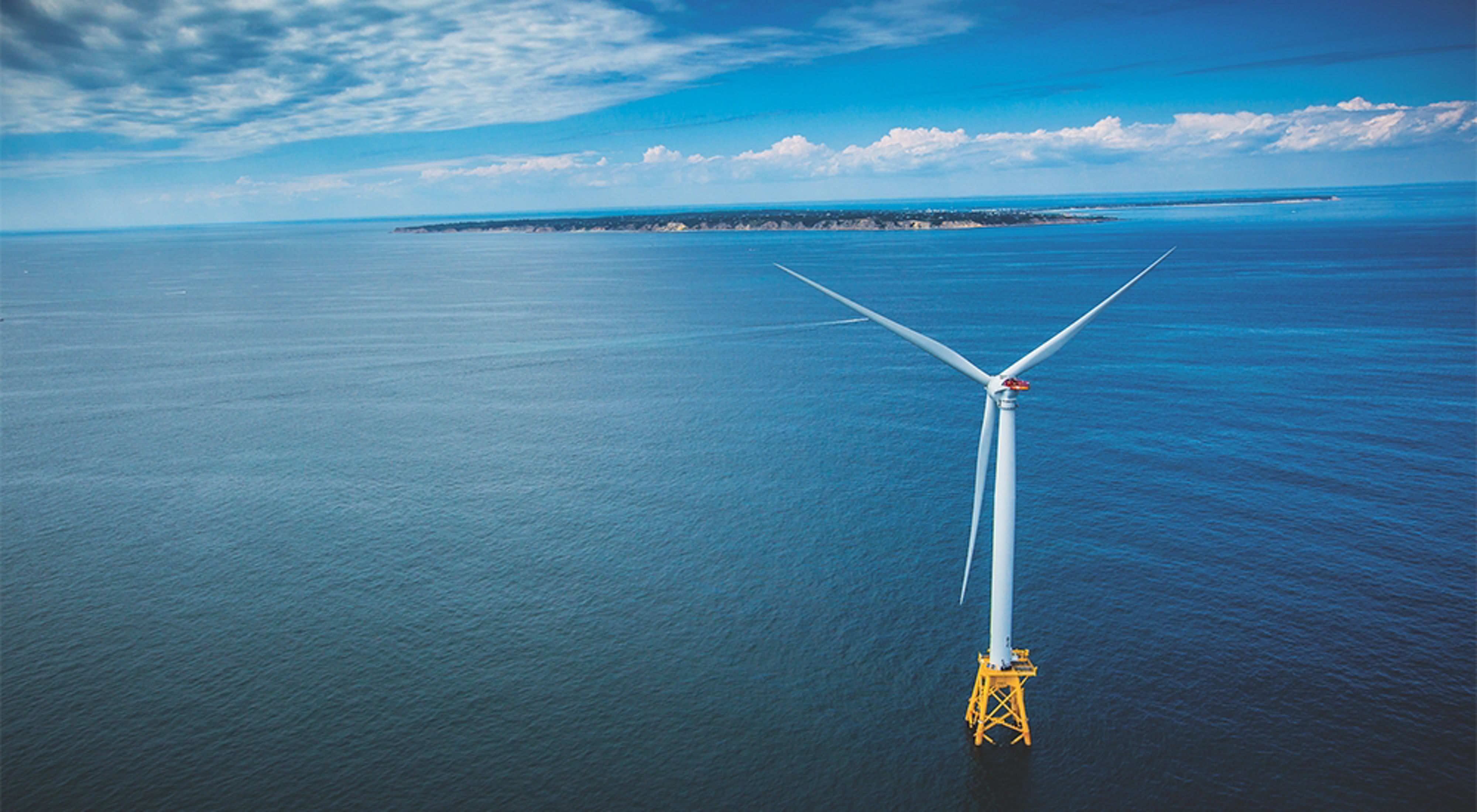 An aerial view of a wind turbine in the ocean with the coast of Block Island and Rhode Island in the background.