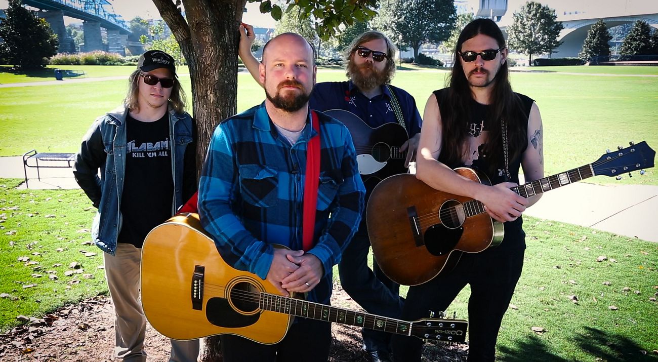 Four musicians standing in a city park under a tree. Three of them are holding guitars.