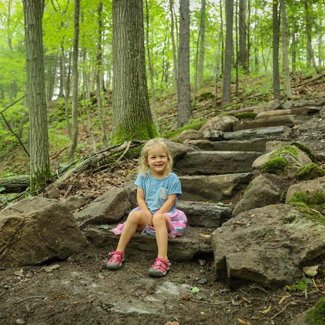 A small child smiles and sits on the bottom step of a rock stairway in the woods.
