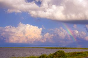 A rainbow peeks through fluffy white clouds above a marsh and Prime Hook Wildlife Refuge. 