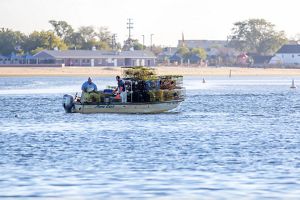 A small motorized boat is shown on calm bay water carrying two people and a large load of stacked wire traps. 