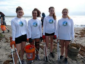 Four volunteers stand with buckets and grabbers in hand to participate in a beach clean-up.