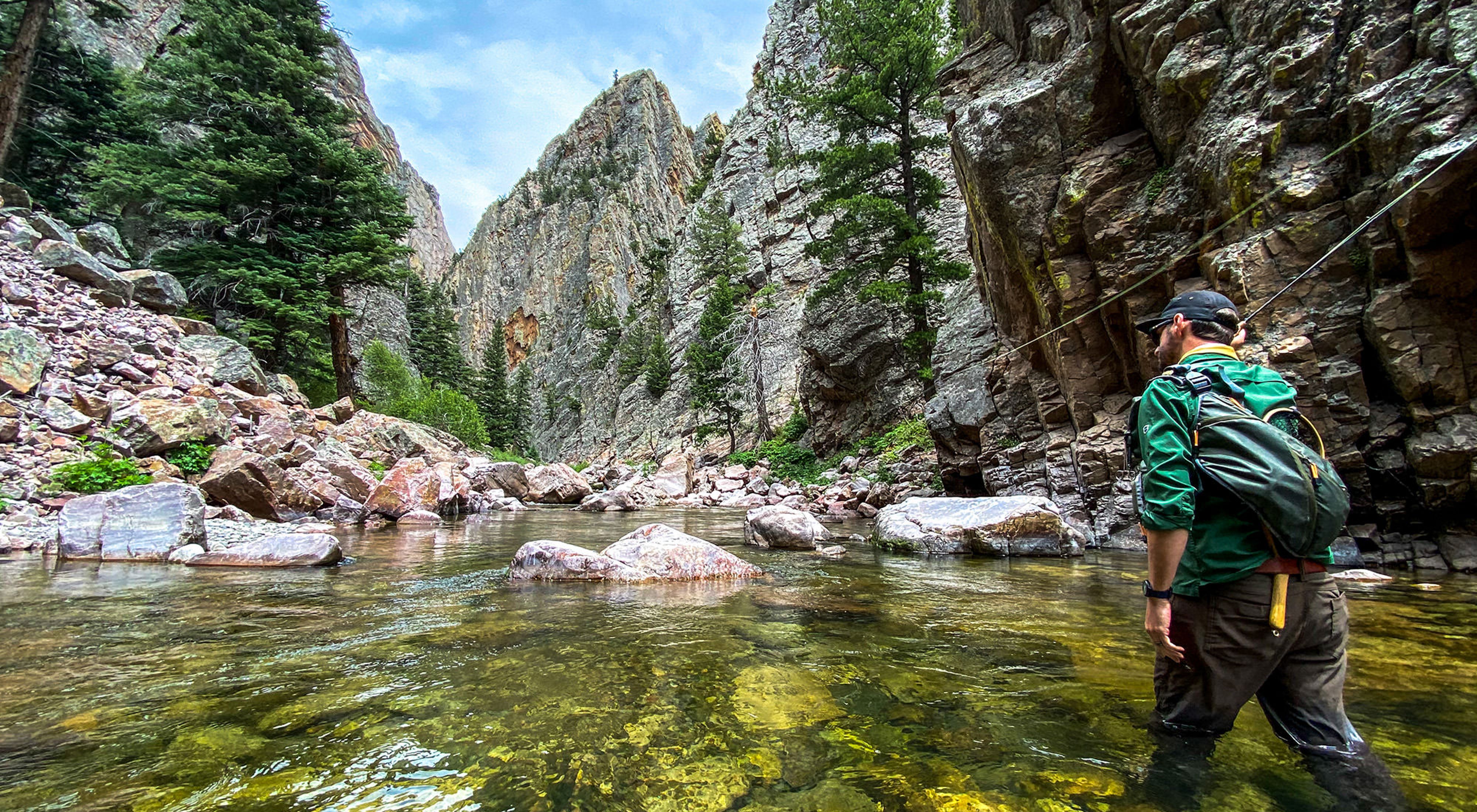 A man wearing a backpack wades through clear water in a rocky canyon.
