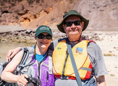 A man and a woman stand with their arms around each other, both wearing hats, sunglasses, and life vests., with water and rocks in the background.