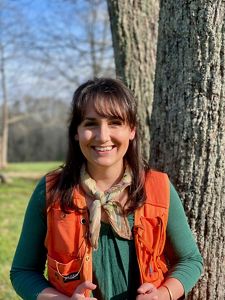 A woman in an orange vest stands in front of a tree.
