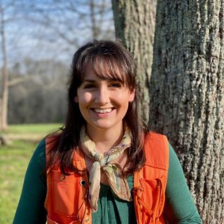 A woman in an orange vest stands in front of a tree.