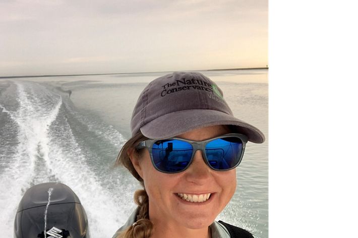 Candid snapshot of Marine Restoration Specialist Brittany Gonzalez. A smiling woman wearing sunglasses and a ball cap drives a small boat in open water.