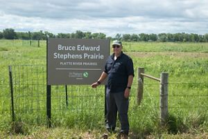 A man stands in front of a new sign that reads Bruce Edward Stephens Prairie Platte River Prairies. 