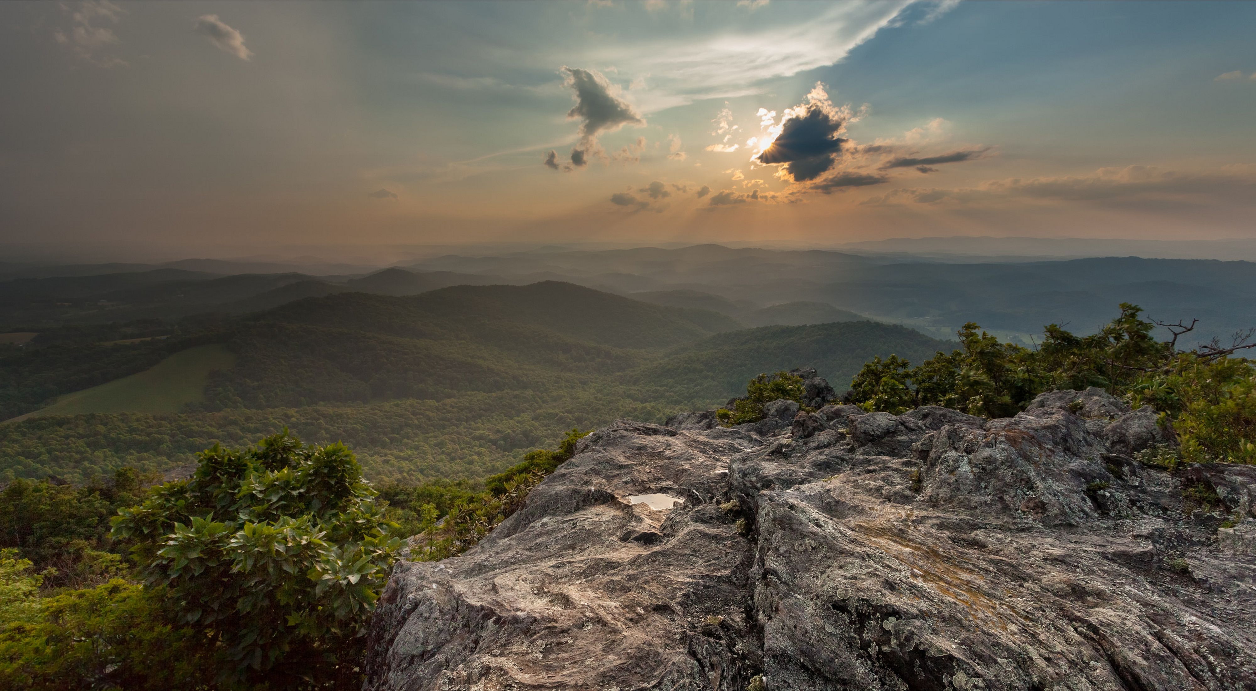 View from a rock outcrop looking over rolling forested green mountain ridge tops as the sun sets behind a bank of clouds.
