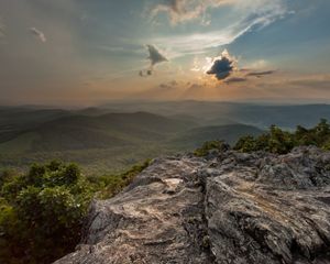 View from a rock outcrop looking over rolling forested green mountain ridge tops as the sun sets behind a bank of clouds.