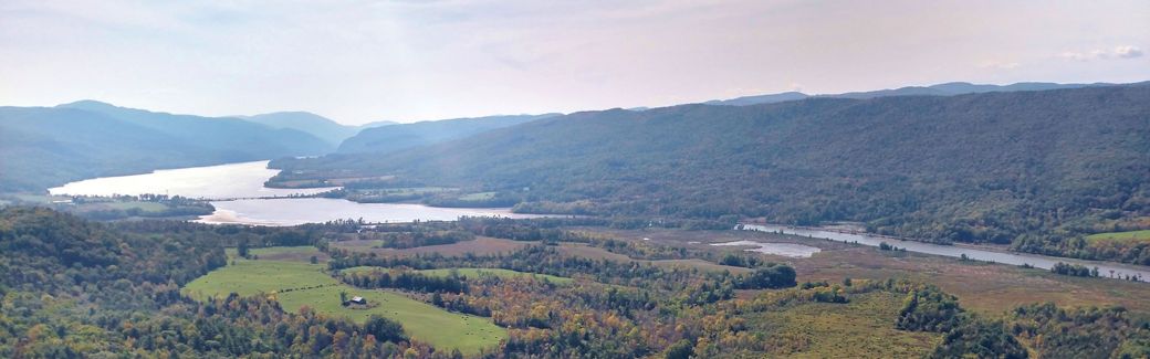 A river flows alongside a valley with green fields and autumn forests, ringed by hills that line the river in the distance.