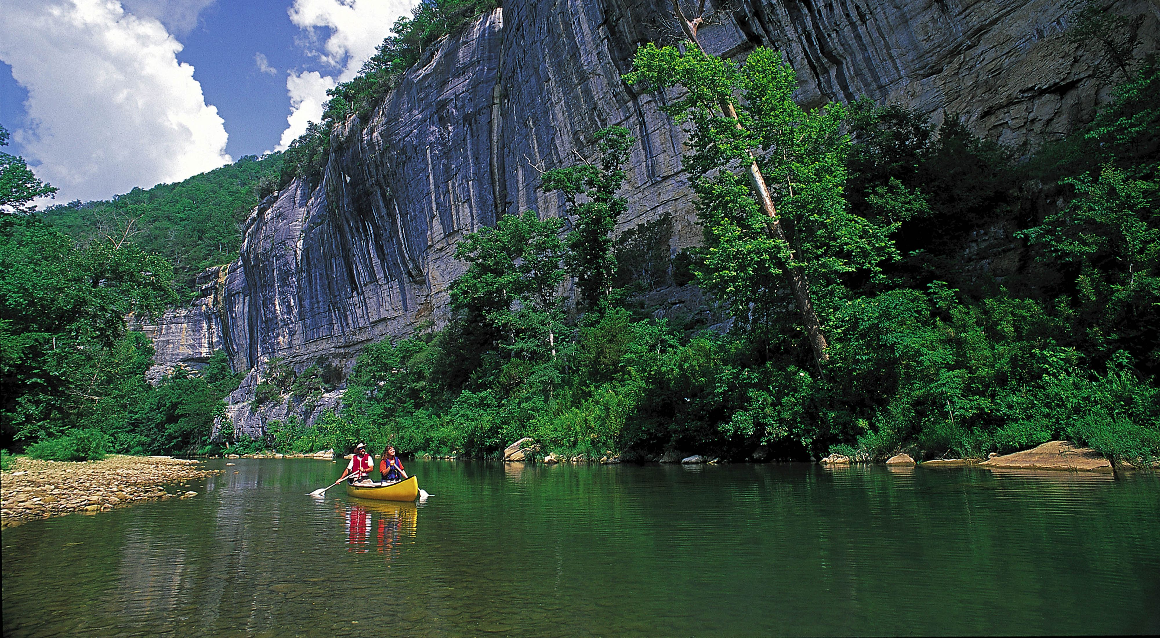 Two people canoeing on the Buffalo River with rocky cliffs along one bank.