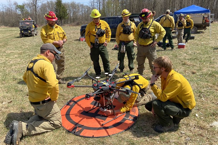Several people in yellow fire gear gather around a drone on the ground.