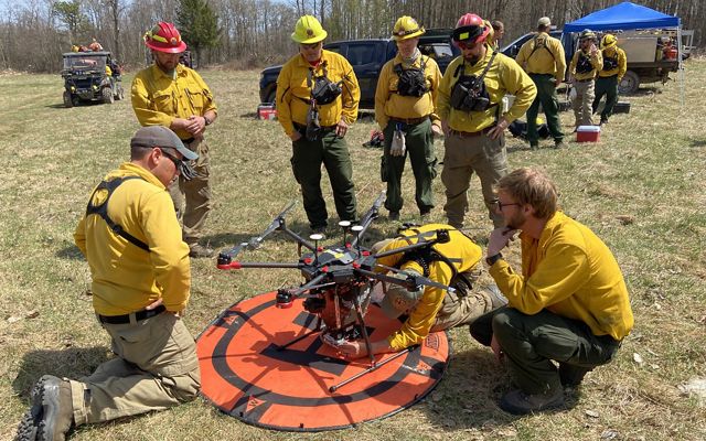 Several people in yellow fire gear gather around a drone on the ground.
