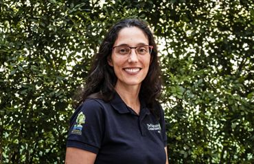 Retrato de Adriana Kfouri de pé em frente a uma folhagem densa.