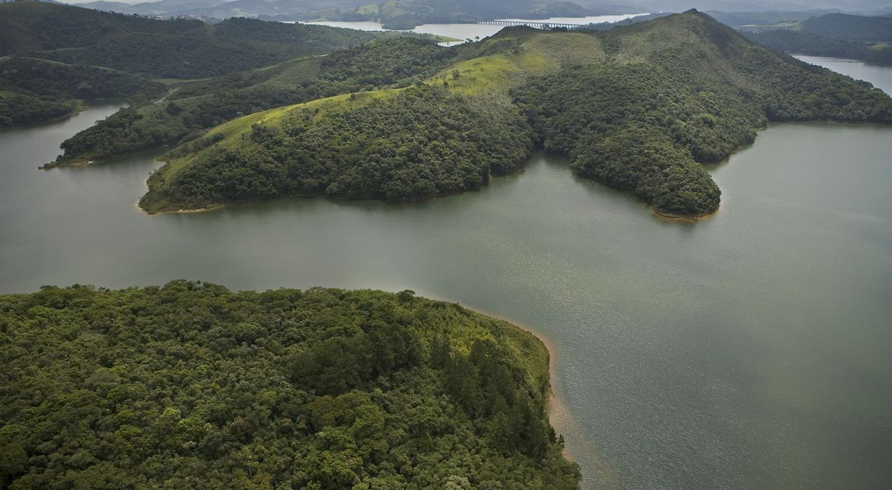 A imagem mostra uma vista aérea de um lago cercado por colinas verdes e floresta densa. Há várias penínsulas se estendendo na água, e ao fundo aparecem mais colinas e montanhas sob um céu nublado.
