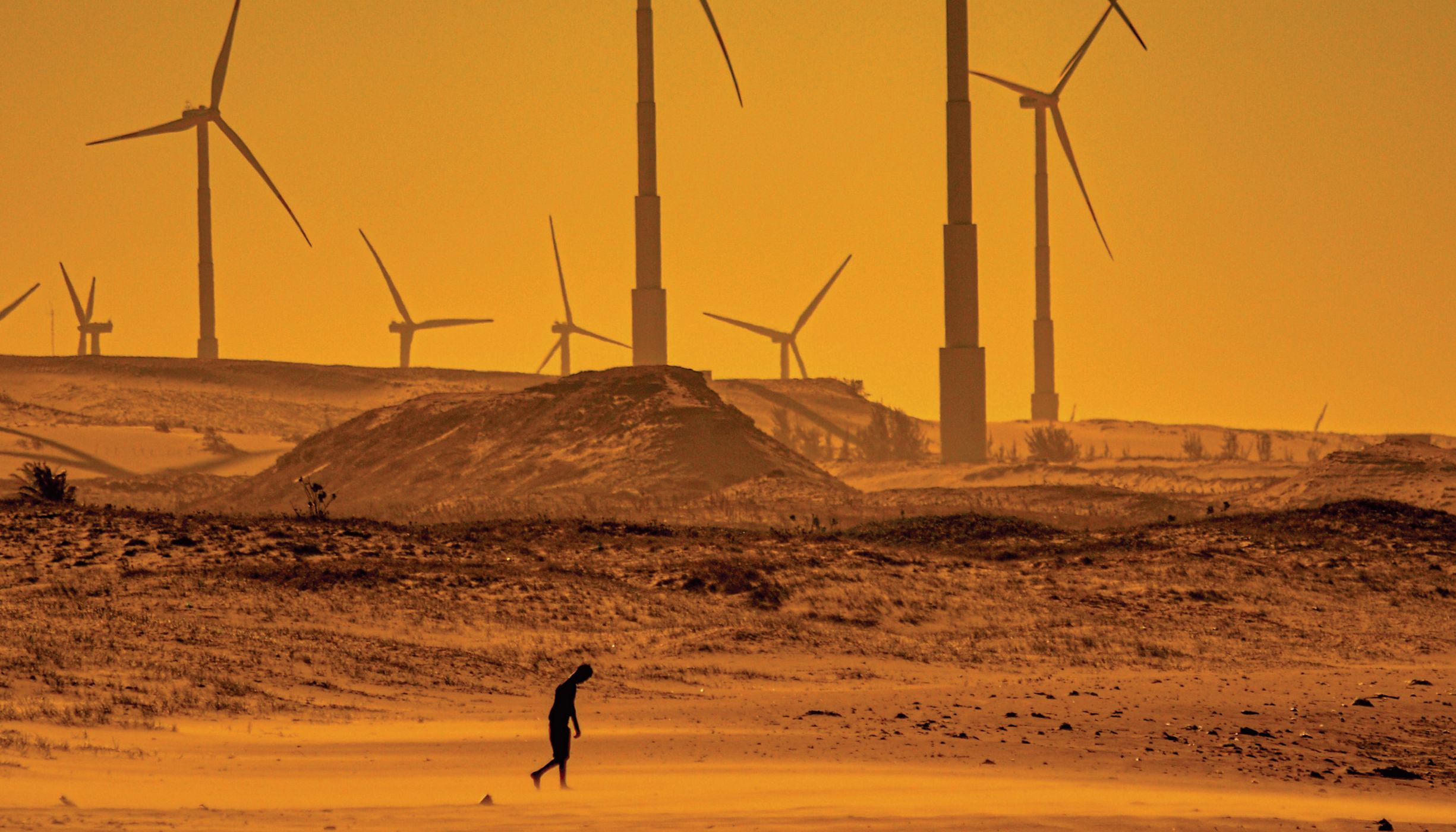 Pessoa andando em paisagem desértica a frente de geradores de energia eólica.