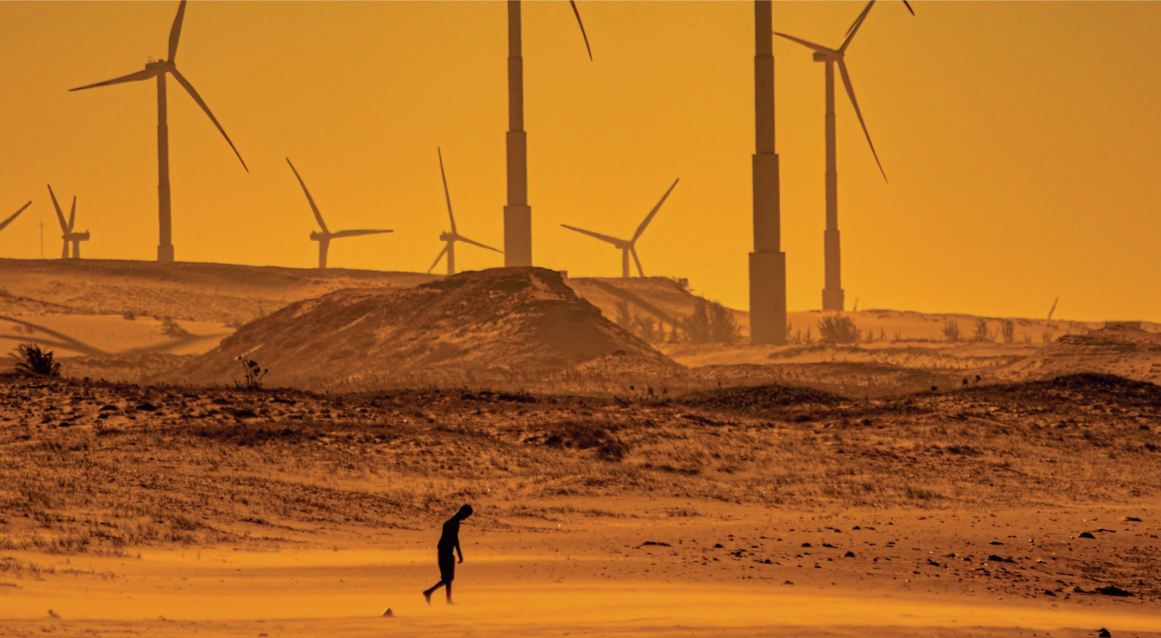 Person walking in desert landscape in front of wind power generators.
