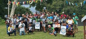 Grande grupo de pessoas posando ao ar livre segurando certificados, com decoração de bandeirinhas e rio ao fundo.