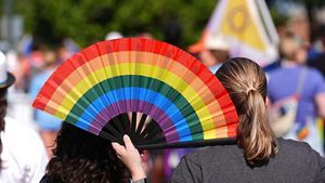 Pessoa segura leque nas cores do arco-íris em evento ao ar livre, com multidão ao fundo, simbolizando orgulho LGBTQIA+ e expressão cultural coletiva.