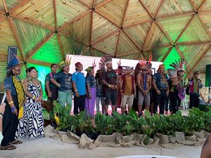   A imagem mostra um grupo de indígenas reunidos em um palco, dentro de uma estrutura de madeira. Eles usam cocares e adornos tradicionais .
