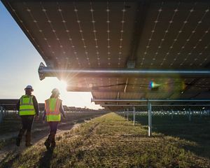 Two people wearing safety vests and hard hats walk alongside an array of solar panels.