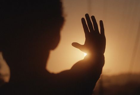 A silhouetted person holds their hand up to block the sun while the sky glows orange.