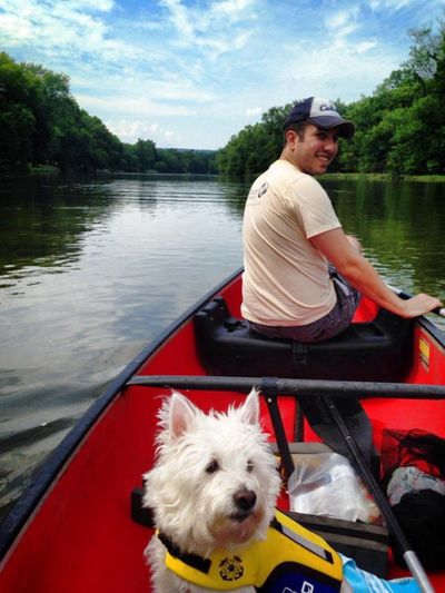 A man sits in the front seat of an orange canoe. He is turned around to look back at the person taking the picture. A small white dog wearing a yellow life vest sits in the middle between them.