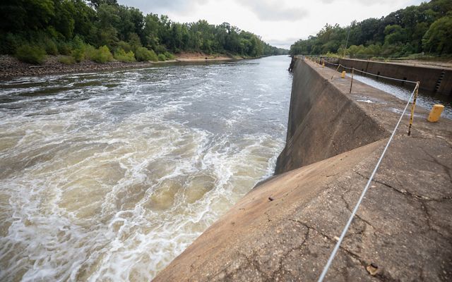 View alongside the wall of a lock and dam with water spilling down in the direction of a river flowing into the distance.