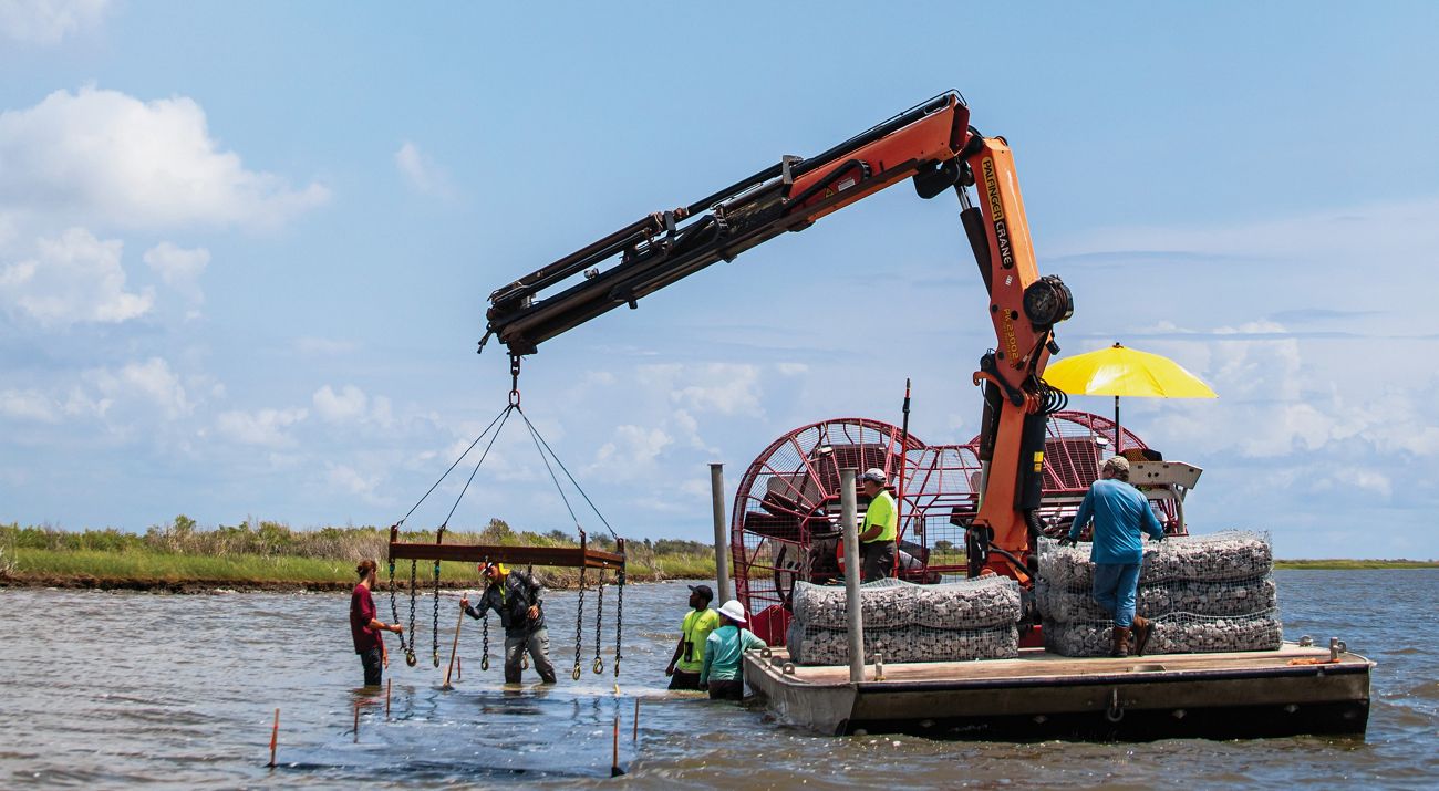 Several people help a flat bottomed boat with a crane through the water.