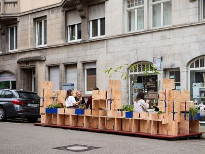 Personas sentadas conversando tras un marco de cajas de madera en las que comienzan a crecer enredaderas.