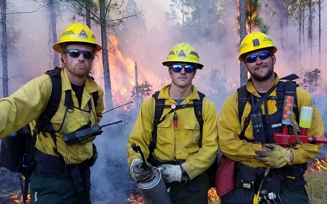 Three students from the University of Idaho dressed in fire-protective gear pose near a prescribed fire at Tiger Creek Preserve. 